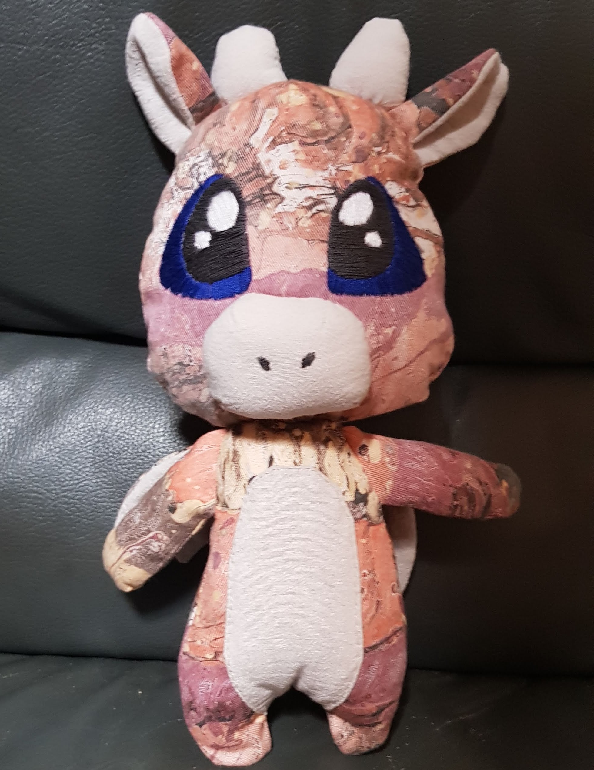 A plush dragon. The main body is made of a canvas like material which has a marbled pattern of oranges, browns and beige. The soft bits, like the inner ear and belly are a plain beige. The dragon has large chibi eyes with which are framed with a vibrant dark blue.