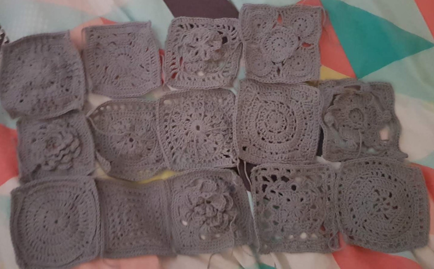 A collection of 14 crocheted granny squares laid flat, crocheted in a gray cotton yarn. Each square is a different pattern with some flat and some 3-d. There are some complex patterns, some simple patterns, and about a third of the patterns are flowers.