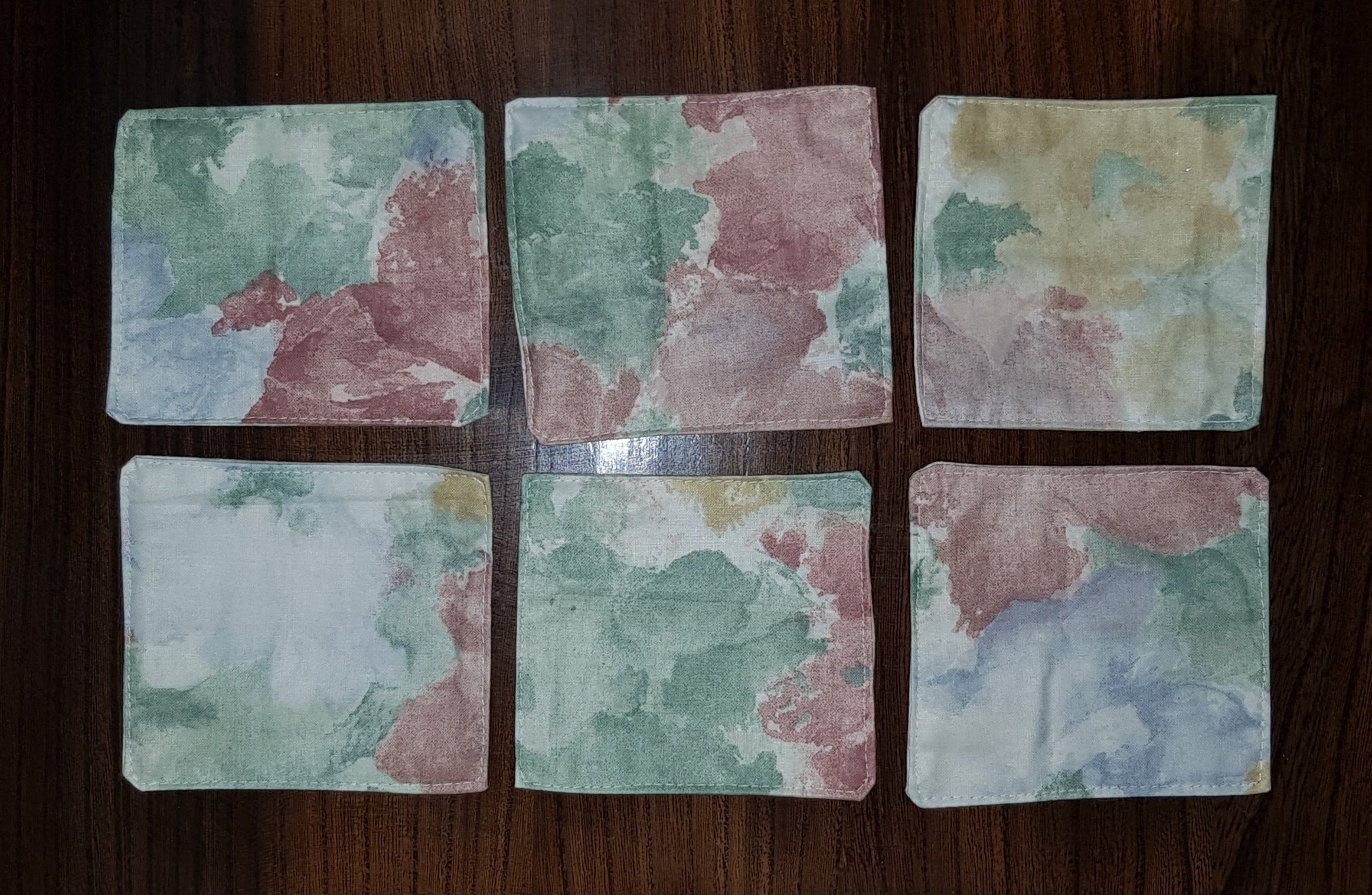 Six fabric coasters sitting on a wooden background. None of the coasters are the same but they have commonalities as they come from the same cloth. The coasters have an off white background with paint-style, matte patches of green, pink, yellow and blue.