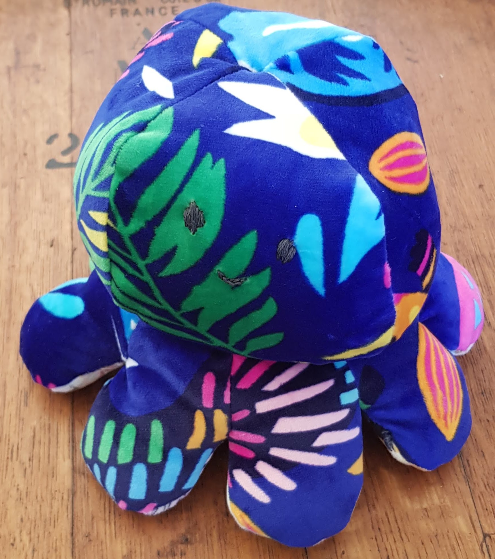 A deep blue plush octopus with multiple colours in many different shapes in the blue; almost jungle-like. The octopus has a happy face.